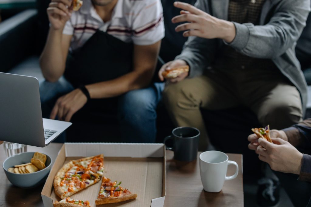 drie mensen zitten achter laptop en eten samen een pizza uit een doos. Personen zijn vanaf schouders in beeld.