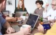 5 mensen aan een tafel naast en tegenover elkaar achter een laptop en met een kop koffie.