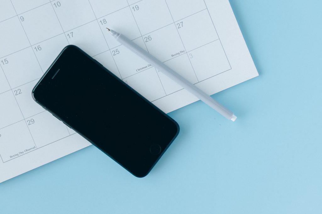 Op een lichtblauwe achtergrond een papieren kalender met een pen en een mobiele telefoon
