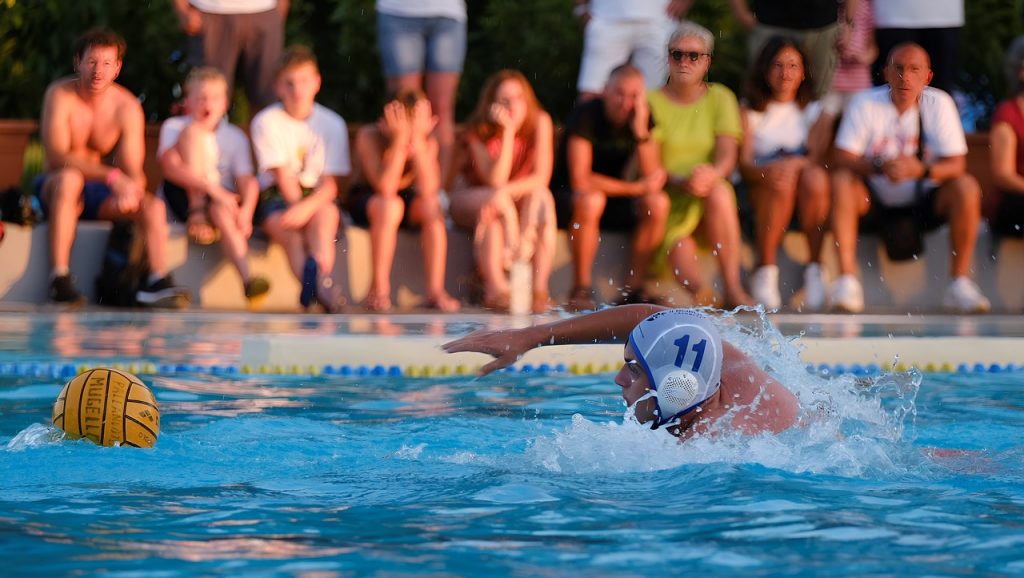 waterpolo speler zwemt naar de bal, op achtergrond publiek