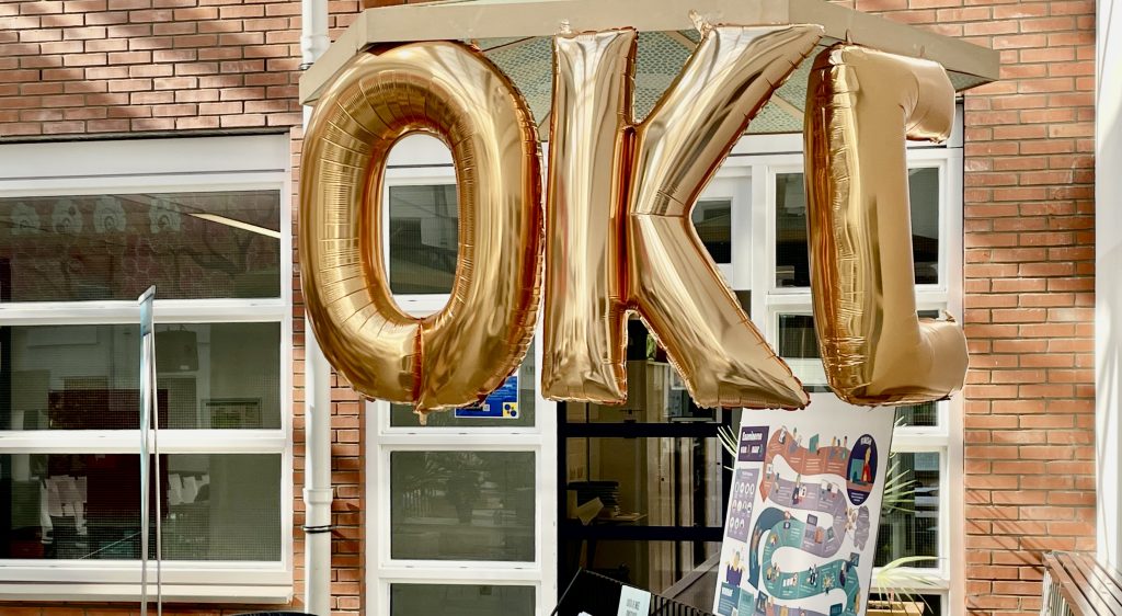 oto van de conferentie met de praatplaten van het OKE project in beeld en drie gouden ballonnen met de letters OKE