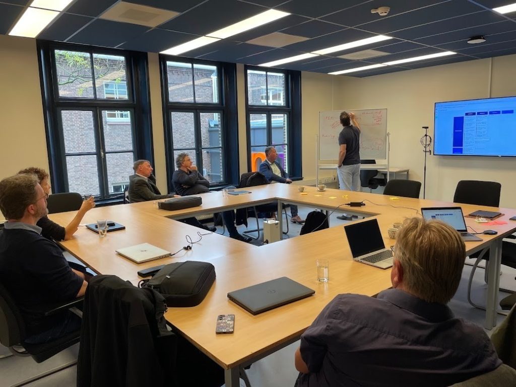 foto van mensen aan tafel in vergaderruimte, kijkend naar iemand die presentatie geeft