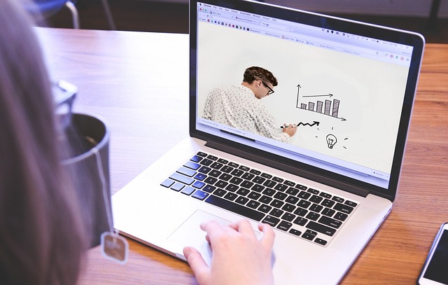 Foto van laptop met daarop beeld van een docent die op een whiteboard met stift grafieken tekent.